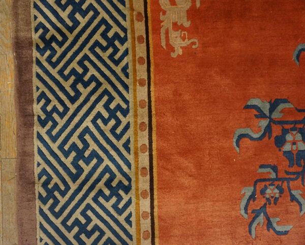 Rust rug with blue botanical patterns and tan geometric key borders resting on light wood flooring.