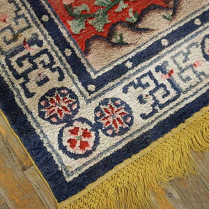 High-pile rug corner showing a red field, navy border, and yellow fringe on distressed wood planks.