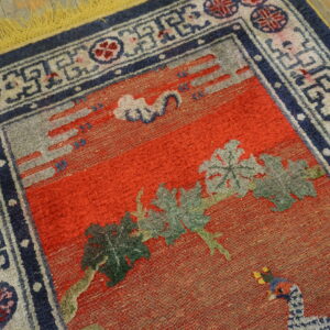 Red field rug with carved green leaves, blue and gray accents, a geometric border, and bright yellow fringe on wood.