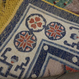 The corner of a patterned rug is turned up, revealing a navy and cream border, red accents, and yellow fringe.
