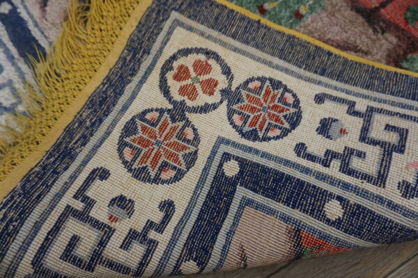 The corner of a patterned rug is turned up, revealing a navy and cream border, red accents, and yellow fringe.