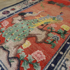 Textured rug detail showing a green bird and floral pattern on a coral red field and wood floor.