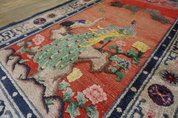 Textured rug detail showing a green bird and floral pattern on a coral red field and wood floor.