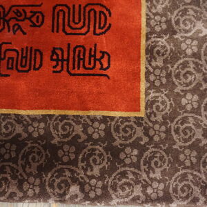 Red rug field with black symbols bordered by dark brown scrolling floral motifs and white fringe.