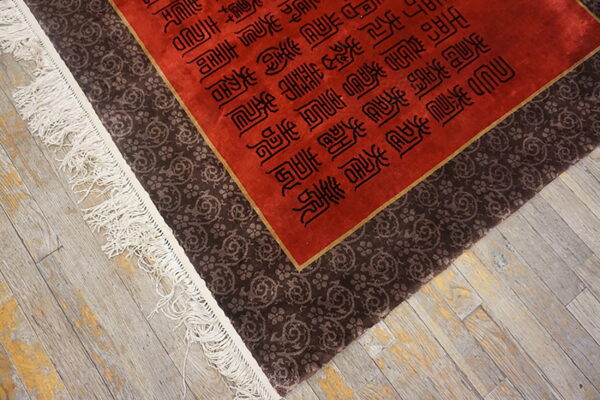 Red rug with black text patterns and a wide brown border rests on distressed grey flooring.