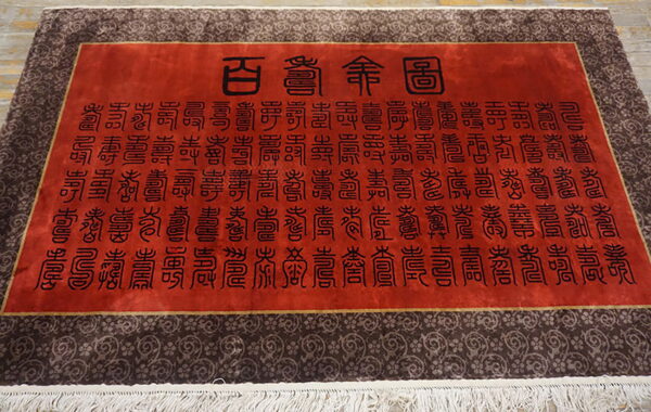 Plush red rug featuring black chinese characters, a thin gold frame, and a wide brown scrolling border.