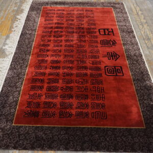 Red rug with black text and a dark brown scrolling border, featuring white fringe on wood flooring.