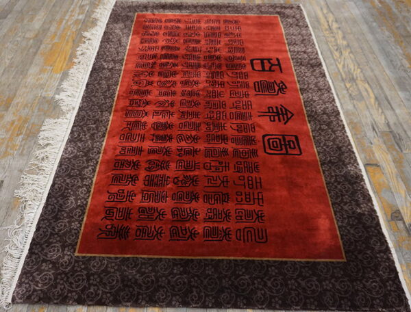 Red rug with black text and a dark brown scrolling border, featuring white fringe on wood flooring.