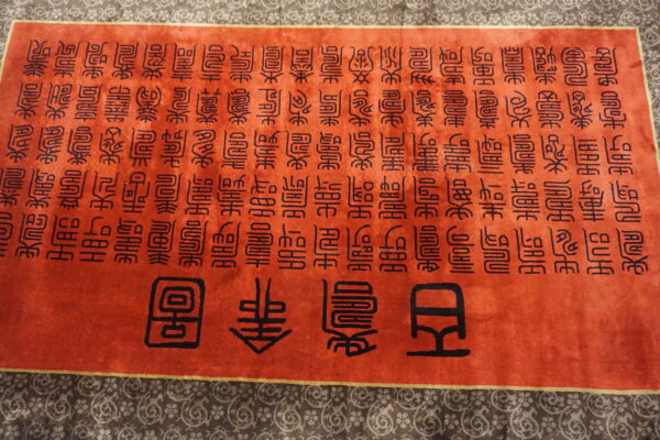 Orange rug displaying black stylized calligraphy covering the field, framed by a patterned outer border.