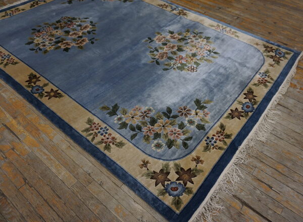 Low-pile blue rug with floral bouquets and a wide tan border lies on rustic, worn hardwood floors.