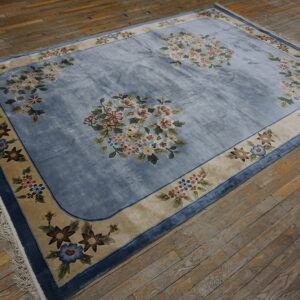 Light blue floral rug with a wide tan border resting on distressed wooden plank flooring.