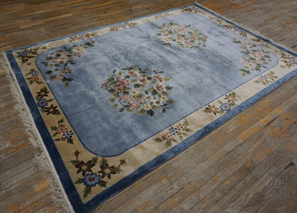 Light blue floral rug with a wide tan border resting on distressed wooden plank flooring.