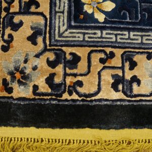 Traditional rug corner in tan, navy, and light blue with a shaggy dark border and yellow fringe on wood.