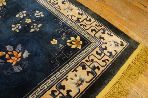 Navy blue rug with floral pattern, cream greek key border, and gold fringe on wood flooring.