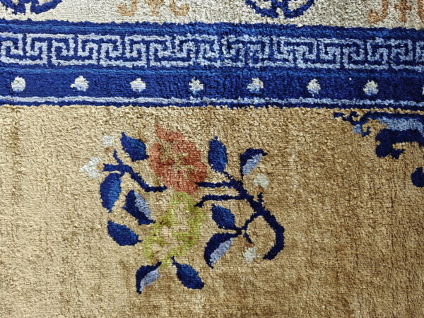 Textured tan rug featuring a blue greek key border and a floral motif in blue, orange, and green.