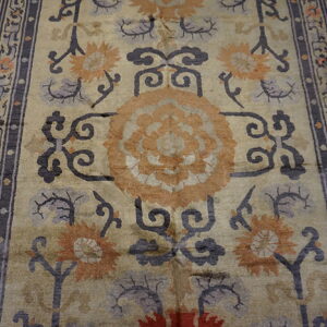 Muted gold rug with stylized orange and gray florals framed by a dark charcoal key pattern border.