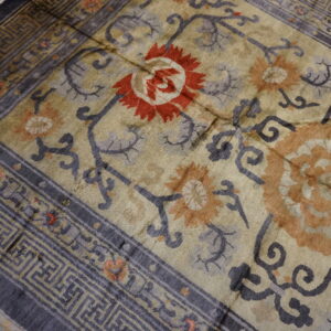 Distressed tan rug with large floral designs, a charcoal border, and geometric fret pattern on wood floor.