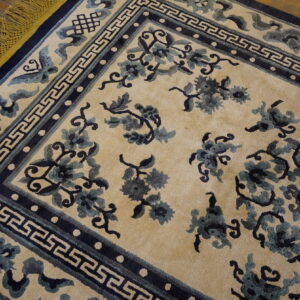 Beige rug with navy and teal floral patterns, geometric border, and mustard yellow fringe on wood floor.