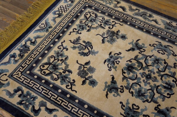 Beige rug with navy and teal floral patterns, geometric border, and mustard yellow fringe on wood floor.