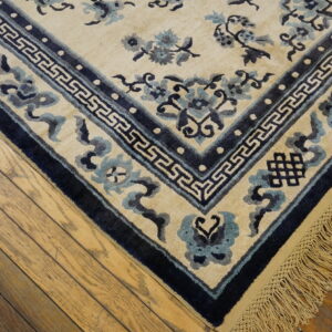 Cream rug with blue floral motifs, geometric border, and fringe on distressed wood floors.