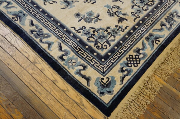 Cream rug with blue floral motifs, geometric border, and fringe on distressed wood floors.