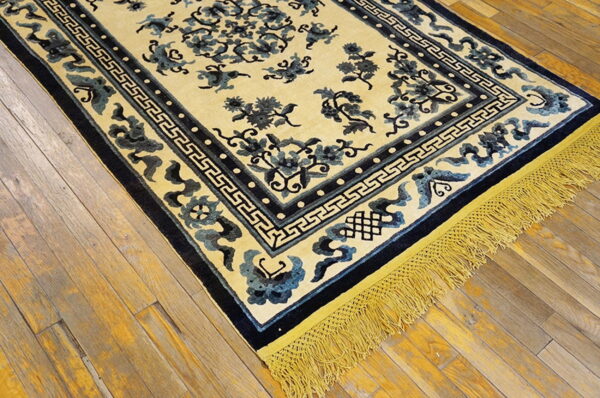 Light yellow rug with dark blue floral patterns, a geometric border, and golden fringe on rustic wood planks.