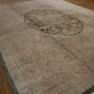 Muted beige rug with sculpted pile, pink accents, and a dark green medallion border sits on wood floors.