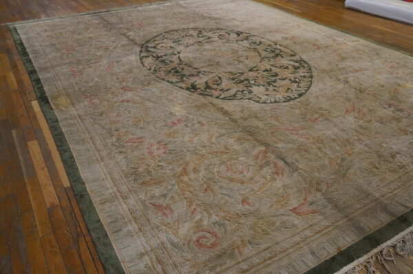Muted beige rug with sculpted pile, pink accents, and a dark green medallion border sits on wood floors.