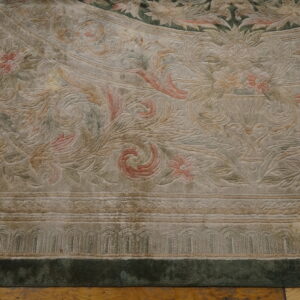 Close-up of a carved rug in light tan, muted pink, and green with scrolling botanical patterns on a wood floor.