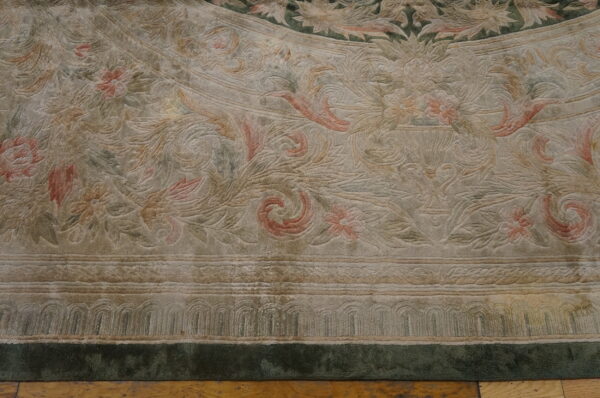 Close-up of a carved rug in light tan, muted pink, and green with scrolling botanical patterns on a wood floor.