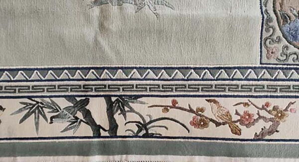 Close-up shows a sage green rug border with embroidered birds, flowers, and geometric greek key trim.