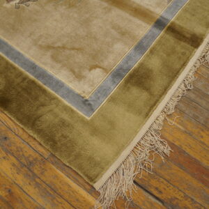 Plush tan rug corner with olive and gray borders and cream fringe on distressed wood flooring.