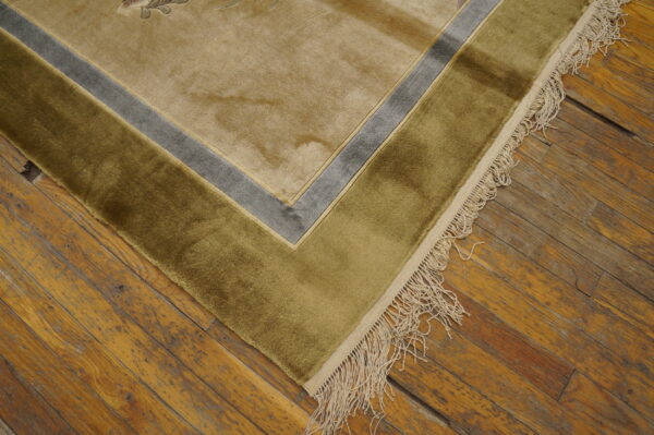 Plush tan rug corner with olive and gray borders and cream fringe on distressed wood flooring.