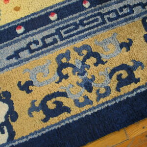A textured rug displays deep blue geometric borders, tan fields, stylized blue patterns, and pink floral accents on wood.