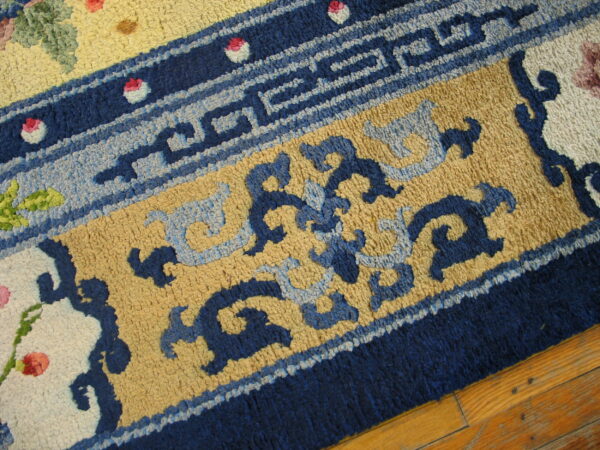 A textured rug displays deep blue geometric borders, tan fields, stylized blue patterns, and pink floral accents on wood.