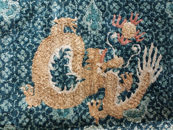 Tan dragon motif with coral accents stands out on a highly textured, mottled teal and blue rug.
