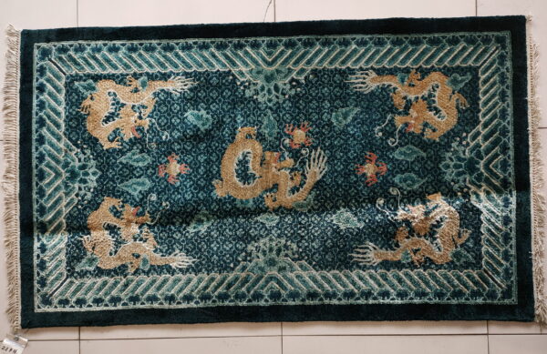 Dark teal high-low pile rug featuring raised gold dragon patterns and a geometric border on white tile.