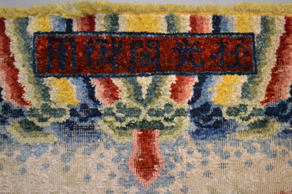Close-up of a high pile rug showing colorful abstract pattern, yellow fringe, and a red field with dark script.
