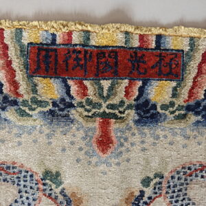 Textured, multicolored rug header showing fringe and a red woven label with blue inscription.