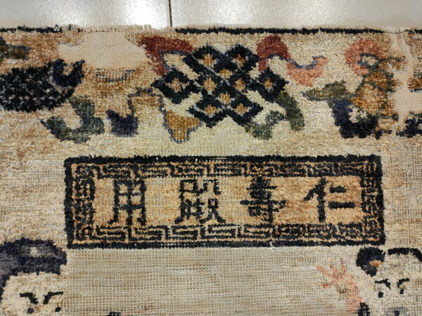 Textured beige rug detail displaying black characters within a rectangular border and a geometric knot motif.