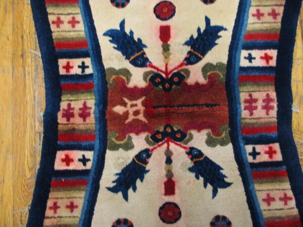 Cream rug detail showing stylized blue fish motifs, rust ground, and multicolored borders on wood plank flooring.