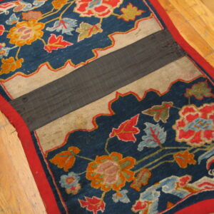 Worn blue rug displaying bright floral patterns, red trim, and a stitched dark gray central seam on light wood floors.