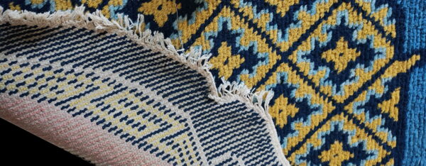 A folded rug shows a navy, gold, and light blue geometric pile pattern next to its woven, striped backing and fringe.