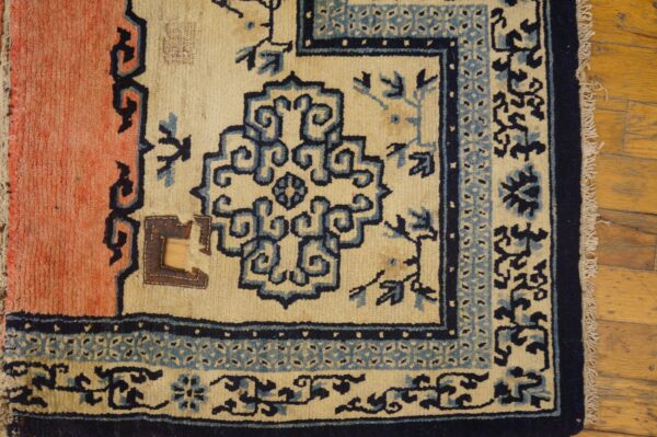 Distressed rug corner featuring cream, coral, and dark blue stylized patterns resting on a light wood floor.