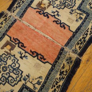 Worn beige, coral, and blue patterned rug with repairs lies on light brown hardwood flooring.