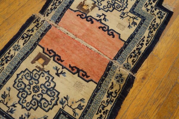 Worn beige, coral, and blue patterned rug with repairs lies on light brown hardwood flooring.