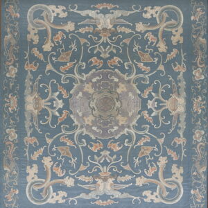 A low-pile rug with a dusty blue field, centered by ornate patterns in beige, gray, and soft peach tones.