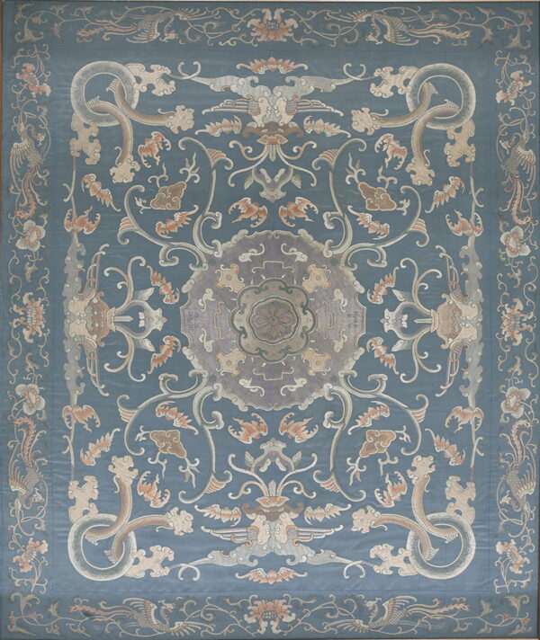 A low-pile rug with a dusty blue field, centered by ornate patterns in beige, gray, and soft peach tones.
