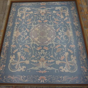 Blue framed textile with beige and terracotta stylized motifs resting on rustic, wide plank wood flooring.