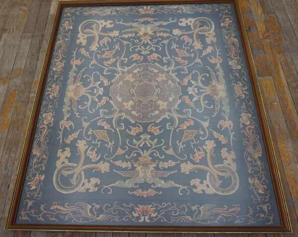 Blue framed textile with beige and terracotta stylized motifs resting on rustic, wide plank wood flooring.